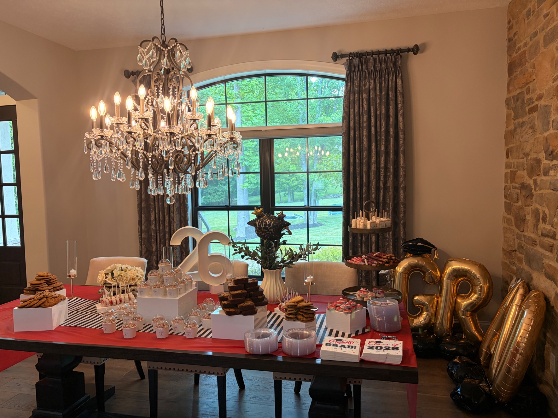 Class of 2026 graduation party dessert table under a crystal chandelier with red linens and gold GRAD balloons