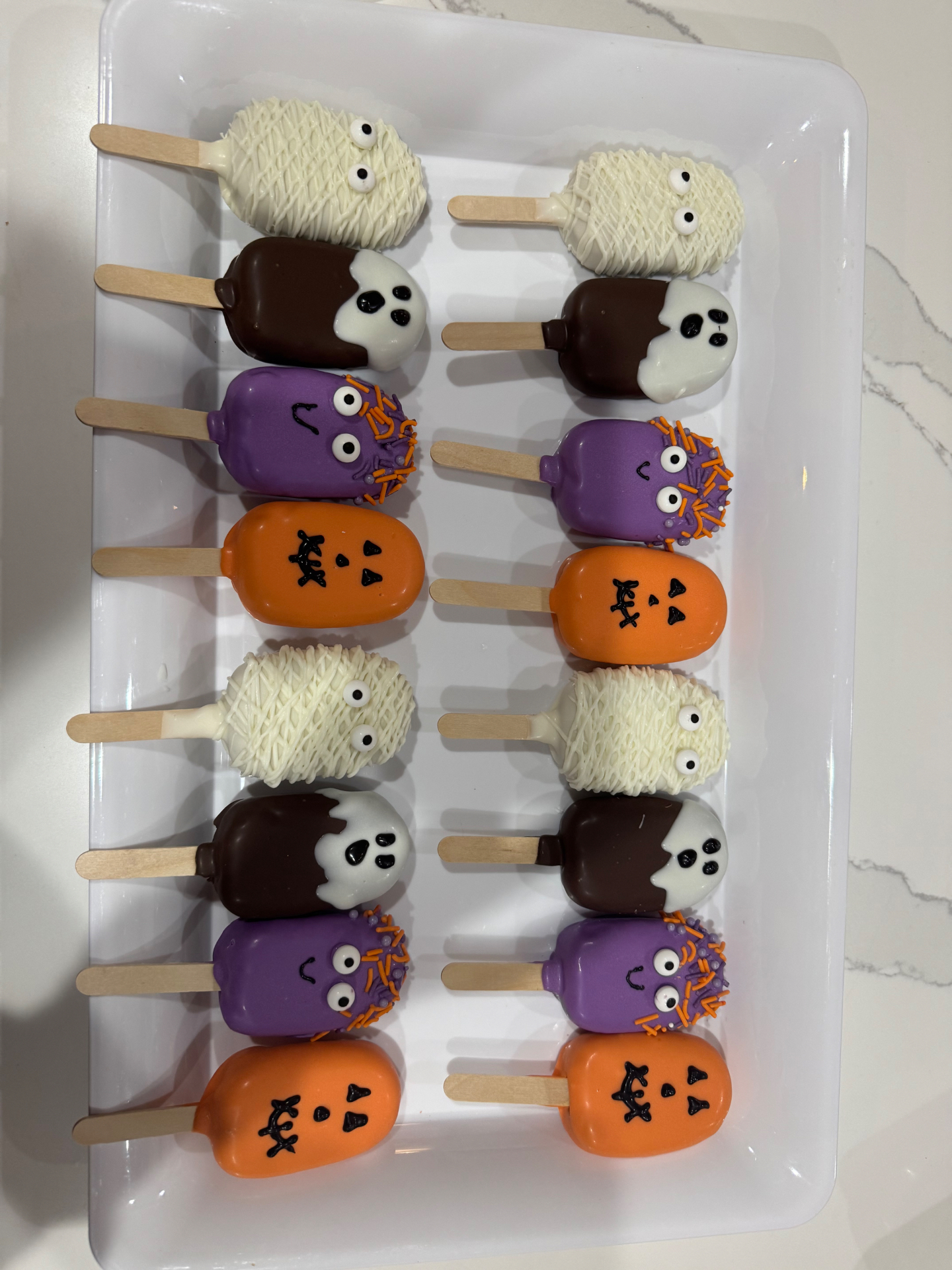 White tray holding sixteen Halloween cake pops arranged in two columns of mummies, ghosts, monsters, and pumpkins