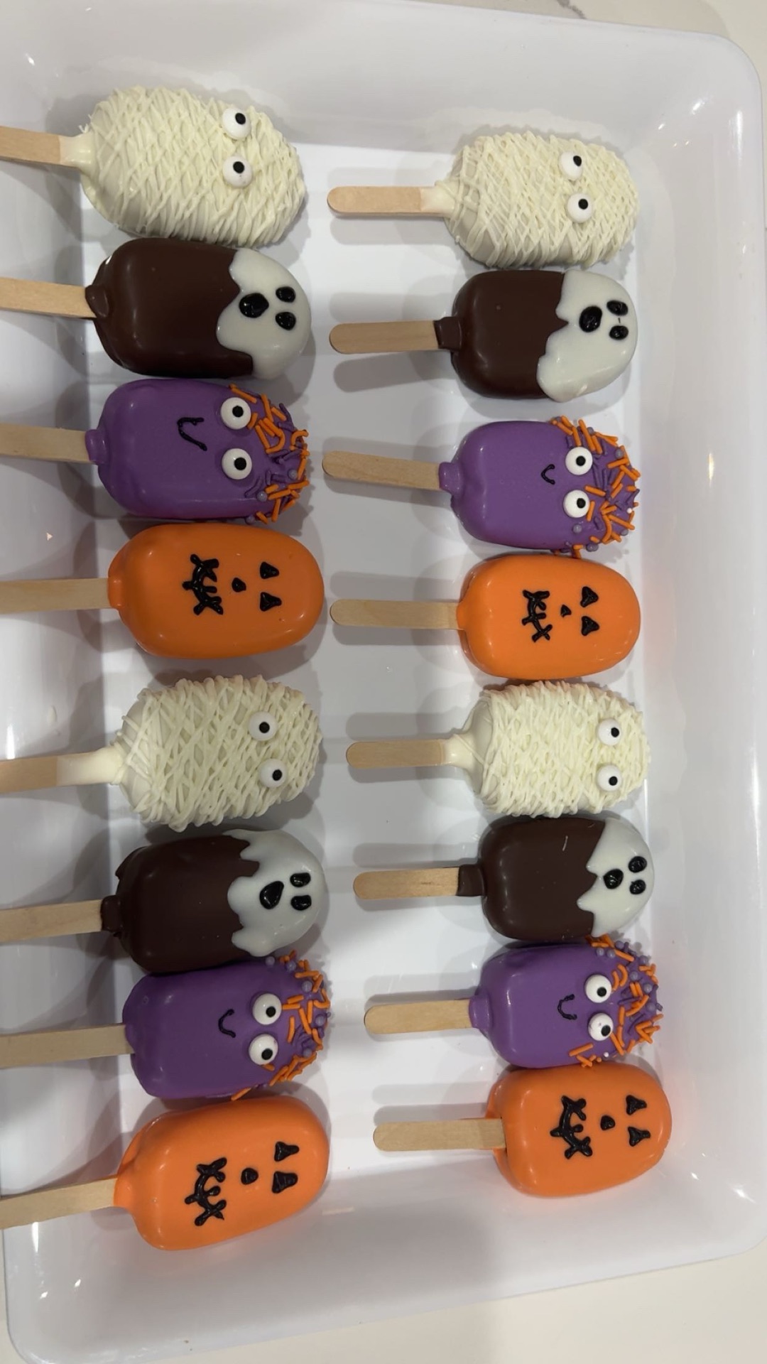 High angle view of a white tray of sixteen Halloween cakesicles in four spooky designs