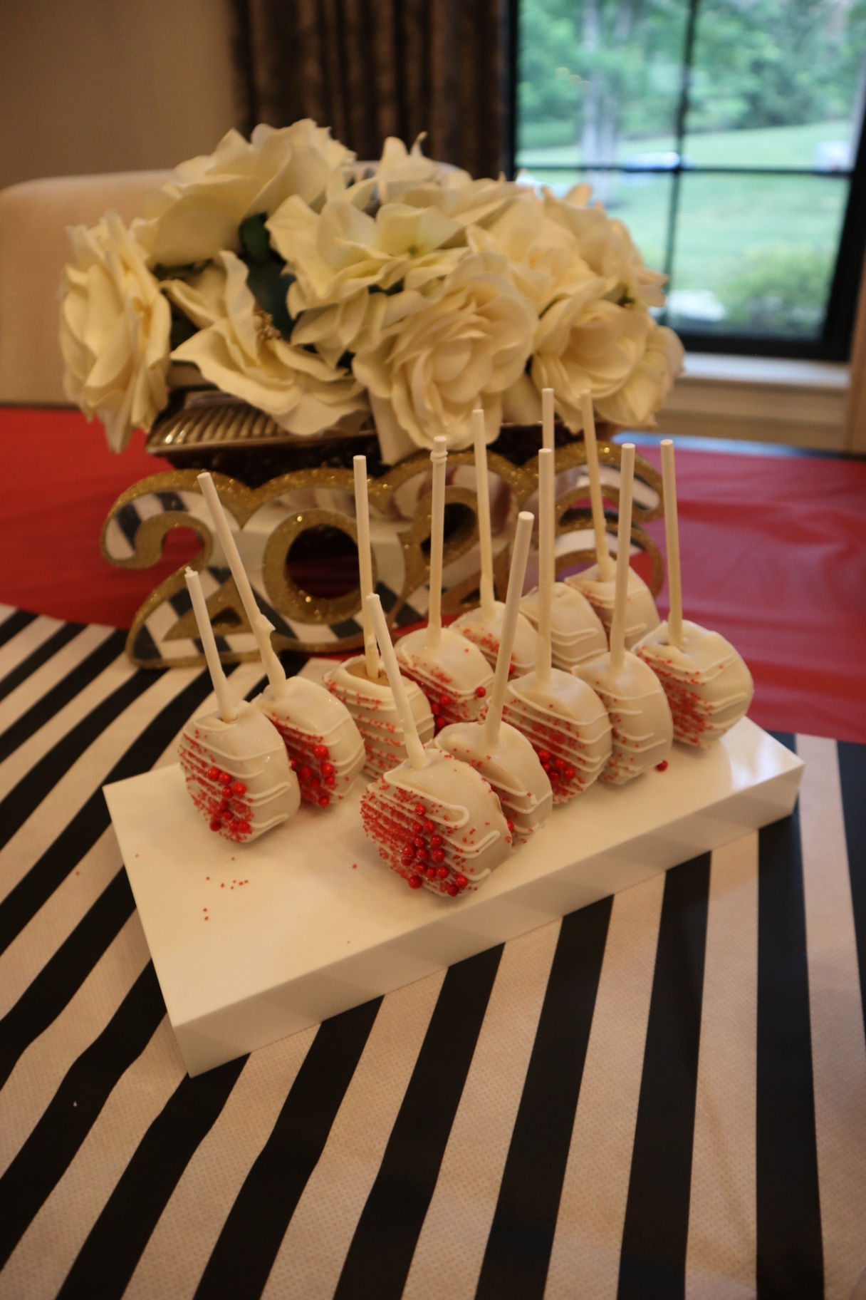 White cake pops with red sprinkles in front of a glitter 2026 sign and white florals