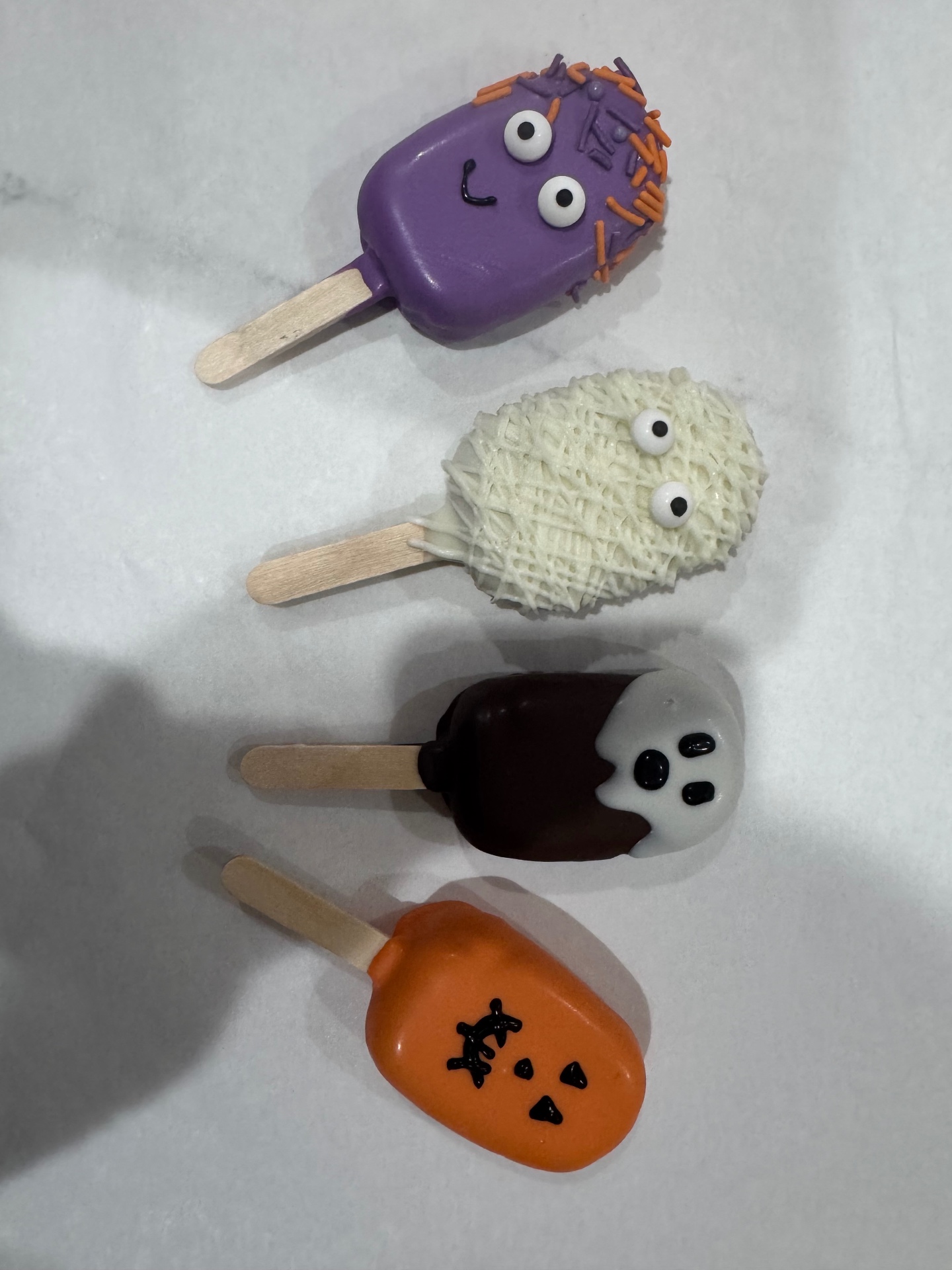 Four Halloween cake pops in a row — a purple monster, a white mummy, a brown ghost, and an orange jack-o'-lantern