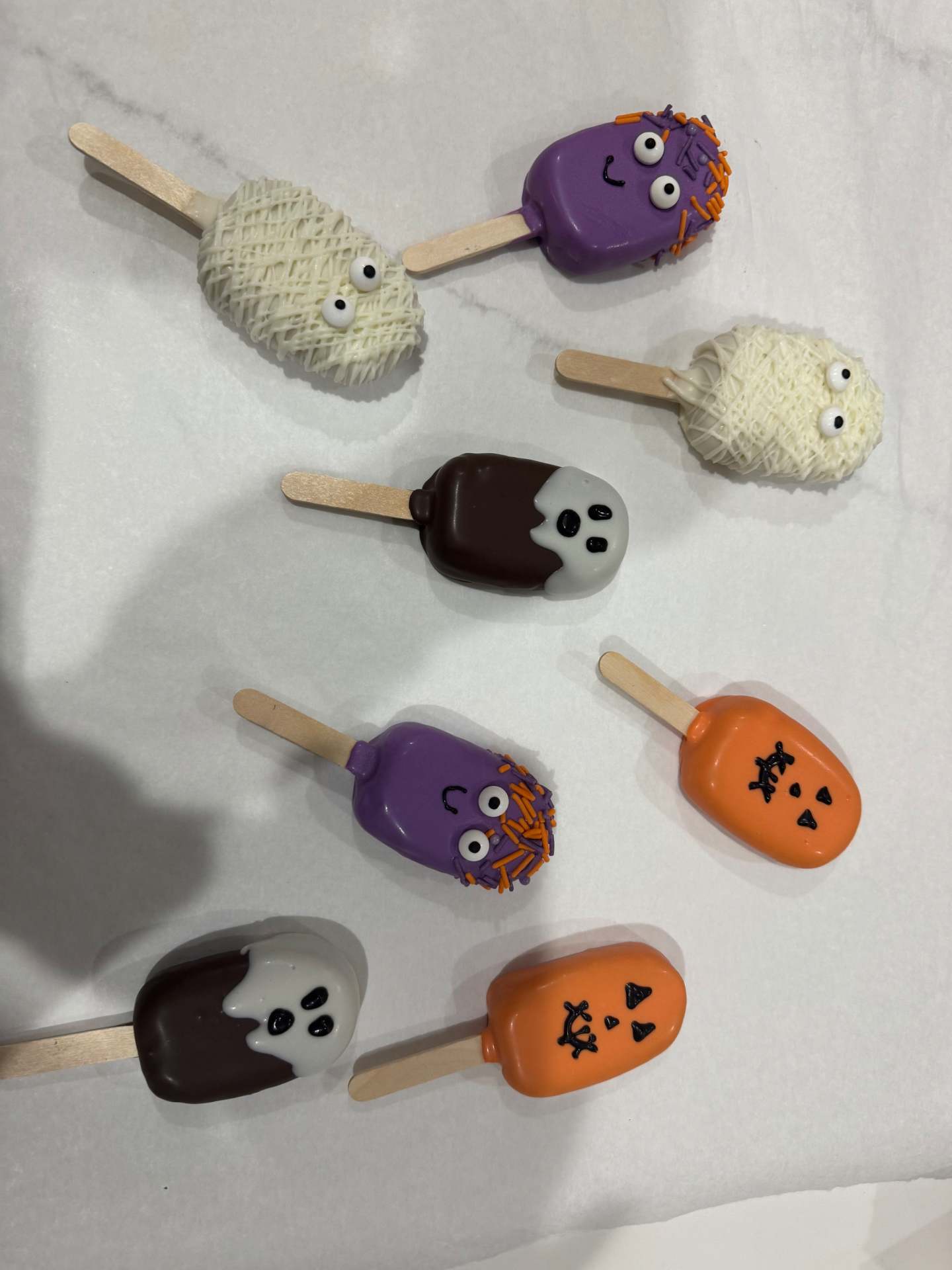 Seven Halloween cakesicles scattered across a white surface — monsters, mummies, ghosts, and a jack-o'-lantern
